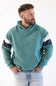 A man wearing a teal colorblock hoodie with navy and white sleeve stripes, paired with light-wash jeans, photographed against a plain white background.