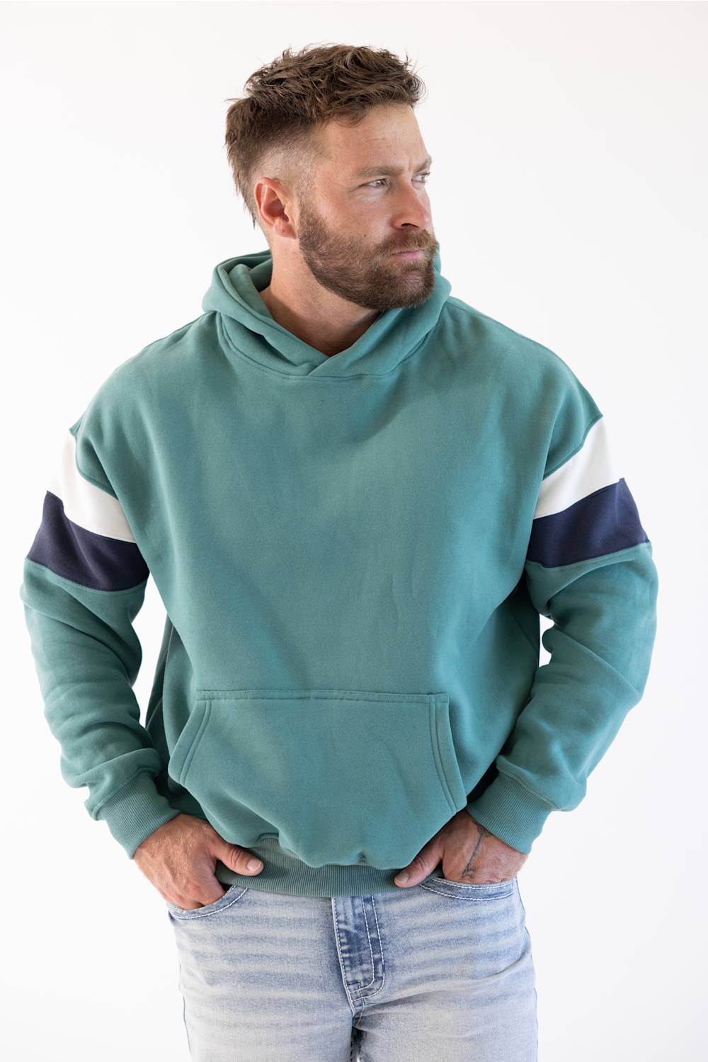 A man wearing a teal colorblock hoodie with navy and white sleeve stripes, paired with light-wash jeans, photographed against a plain white background.
