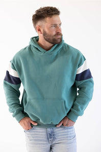 A man wearing a teal colorblock hoodie with navy and white sleeve stripes, paired with light-wash jeans, photographed against a plain white background.