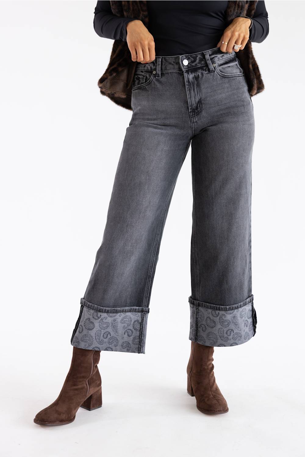 A person wearing dark gray cropped jeans with rolled cuffs showing a paisley pattern, styled with brown ankle boots and a dark jacket against a plain white studio background.