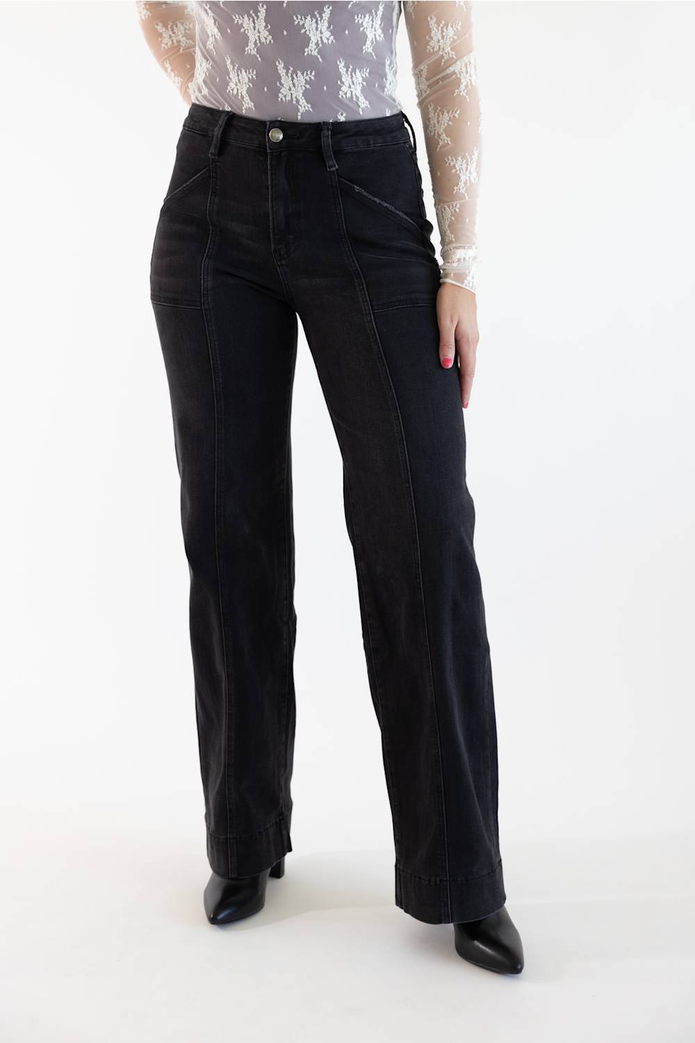 A model wearing a pair of black wide-leg denim jeans, styled with a sheer white patterned long-sleeve top and black pointed-toe heels, against a plain white studio background.