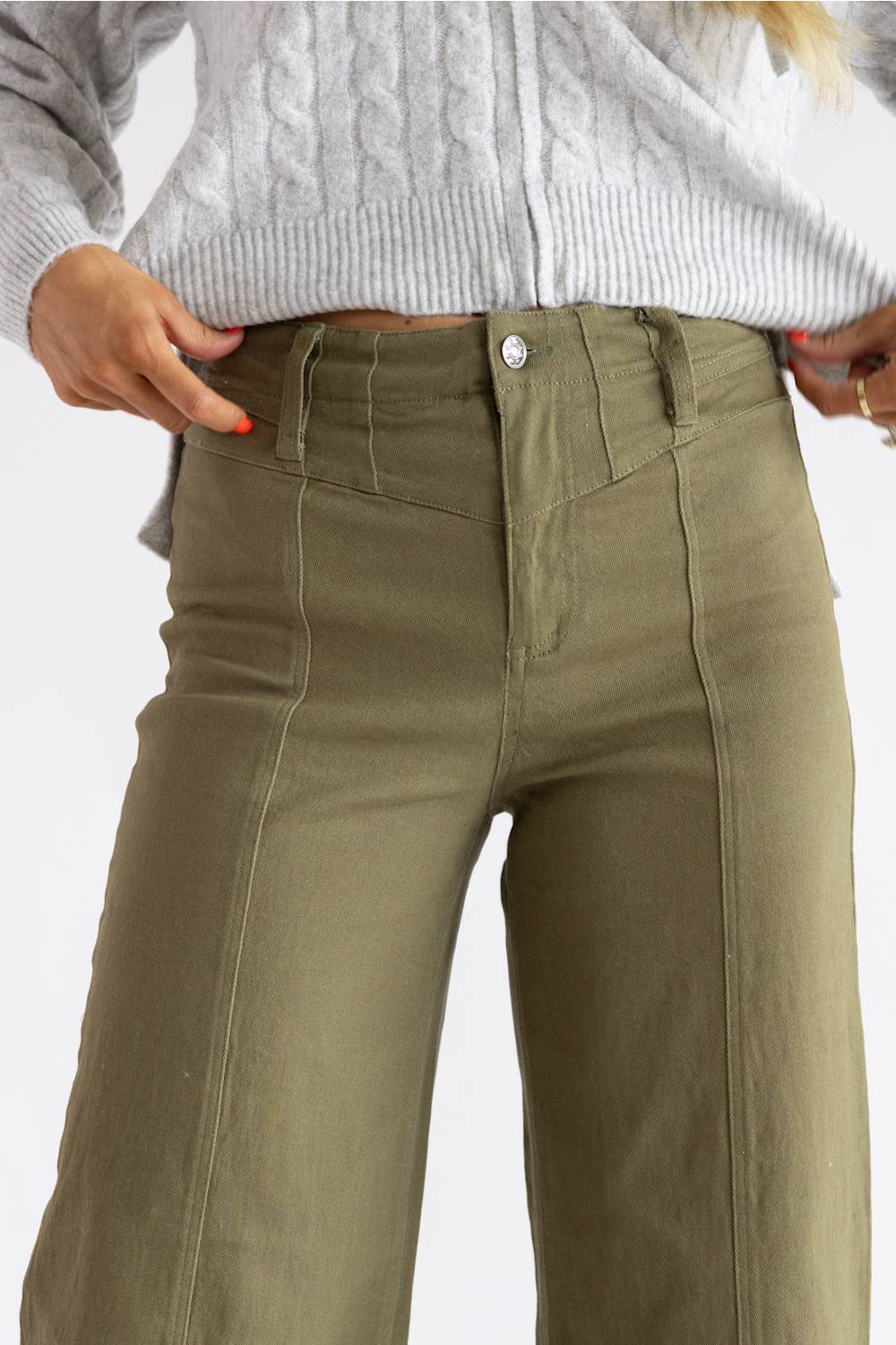 A close-up of a person wearing olive green wide-leg pants with a gray cable-knit sweater, shown from the chest down against a light gray background.
