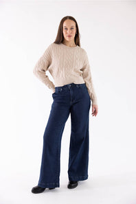 A woman modeling a beige cable-knit sweater and dark blue wide-leg jeans against a plain white studio background.