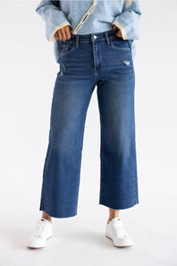 A person wearing blue wide-leg cropped jeans with a light blue fuzzy-sleeve top and white sneakers, standing against a plain white studio background.