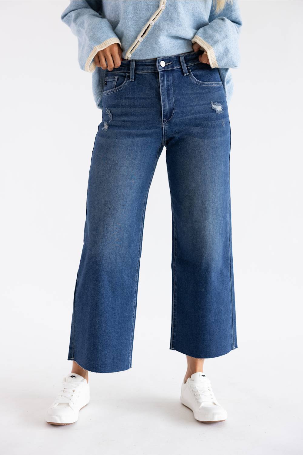 A person wearing blue wide-leg cropped jeans with a light blue fuzzy-sleeve top and white sneakers, standing against a plain white studio background.