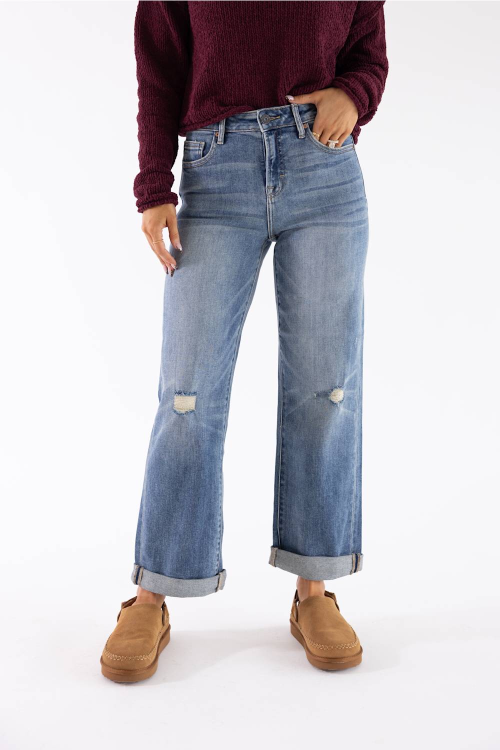 A person wearing light-wash blue wide-leg jeans with distressed knee details and rolled cuffs, paired with a dark burgundy sweater and tan slip-on shoes, against a plain white studio background.
