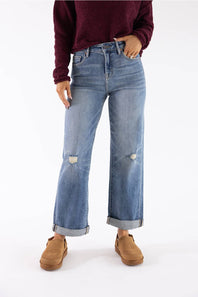 A person wearing light-wash blue wide-leg jeans with distressed knee details and rolled cuffs, paired with a dark burgundy sweater and tan slip-on shoes, against a plain white studio background.