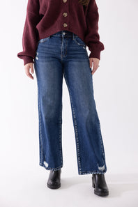 Front close-up view of a model wearing KanCan dark wash, high-rise wide-leg jeans with slight distressing at the hem and thighs, styled with black boots and a button-front burgundy sweater against a plain backdrop.