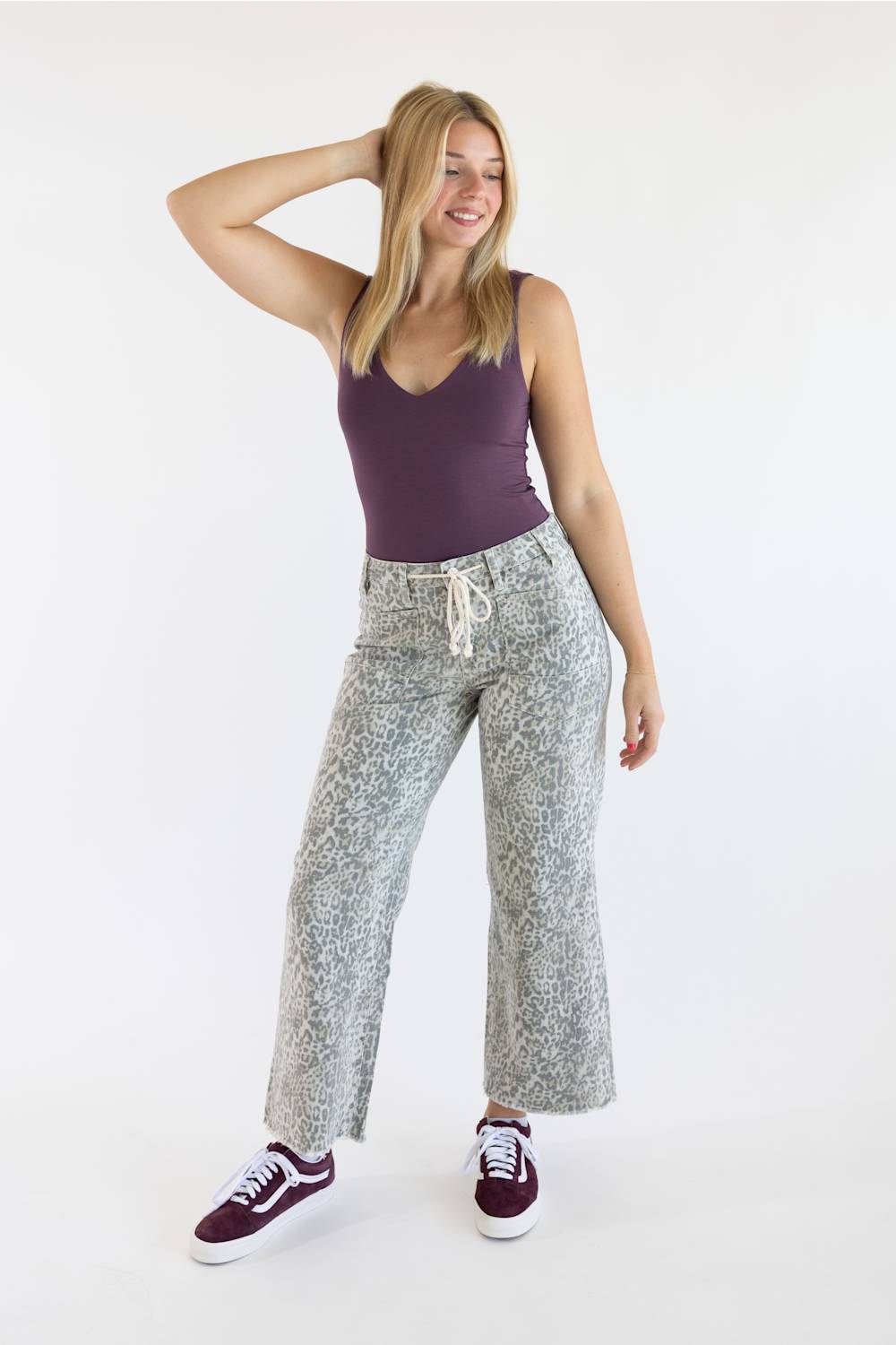 A blonde model wearing a dark purple tank top, light grey leopard print wide-leg pants with a drawstring waist, and maroon sneakers against a white background.
