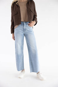 A model wearing light-wash wide-leg blue jeans, styled with a brown cropped jacket, a beige ribbed top, and white sneakers, photographed against a seamless white studio background.