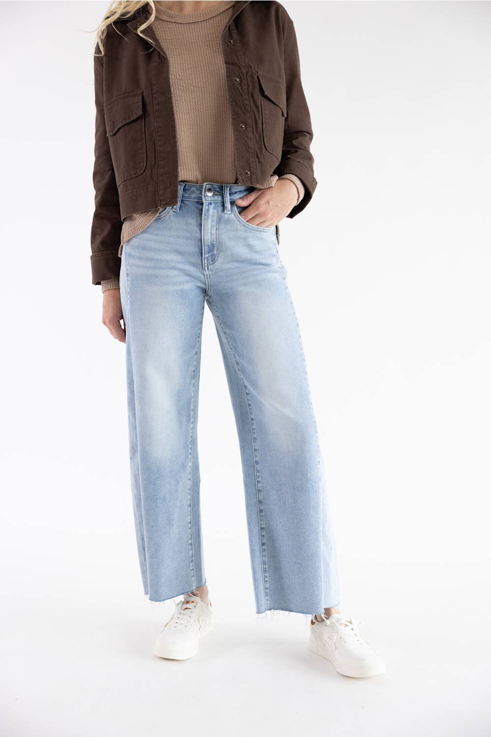 A model wearing light-wash wide-leg blue jeans, styled with a brown cropped jacket, a beige ribbed top, and white sneakers, photographed against a seamless white studio background.