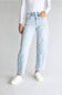 A model wearing light-wash blue high-rise jeans with diagonal patchwork paneling on the front of the legs, styled with a white long-sleeve top and white sneakers against a light gray studio background.
