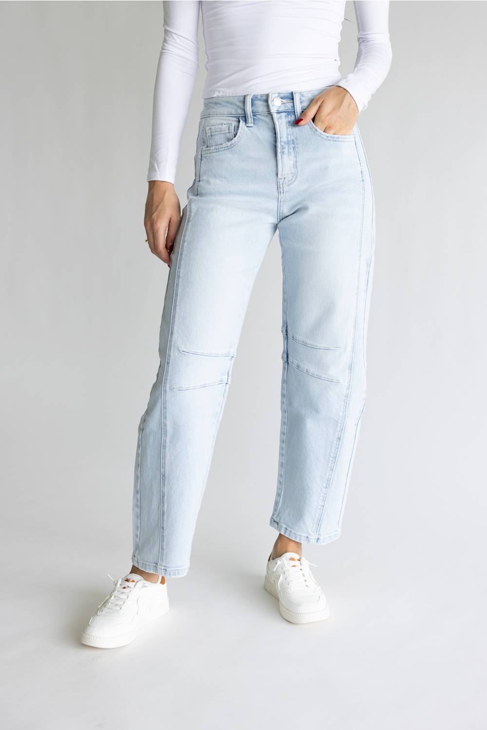 A model wearing light-wash blue high-rise jeans with diagonal patchwork paneling on the front of the legs, styled with a white long-sleeve top and white sneakers against a light gray studio background.