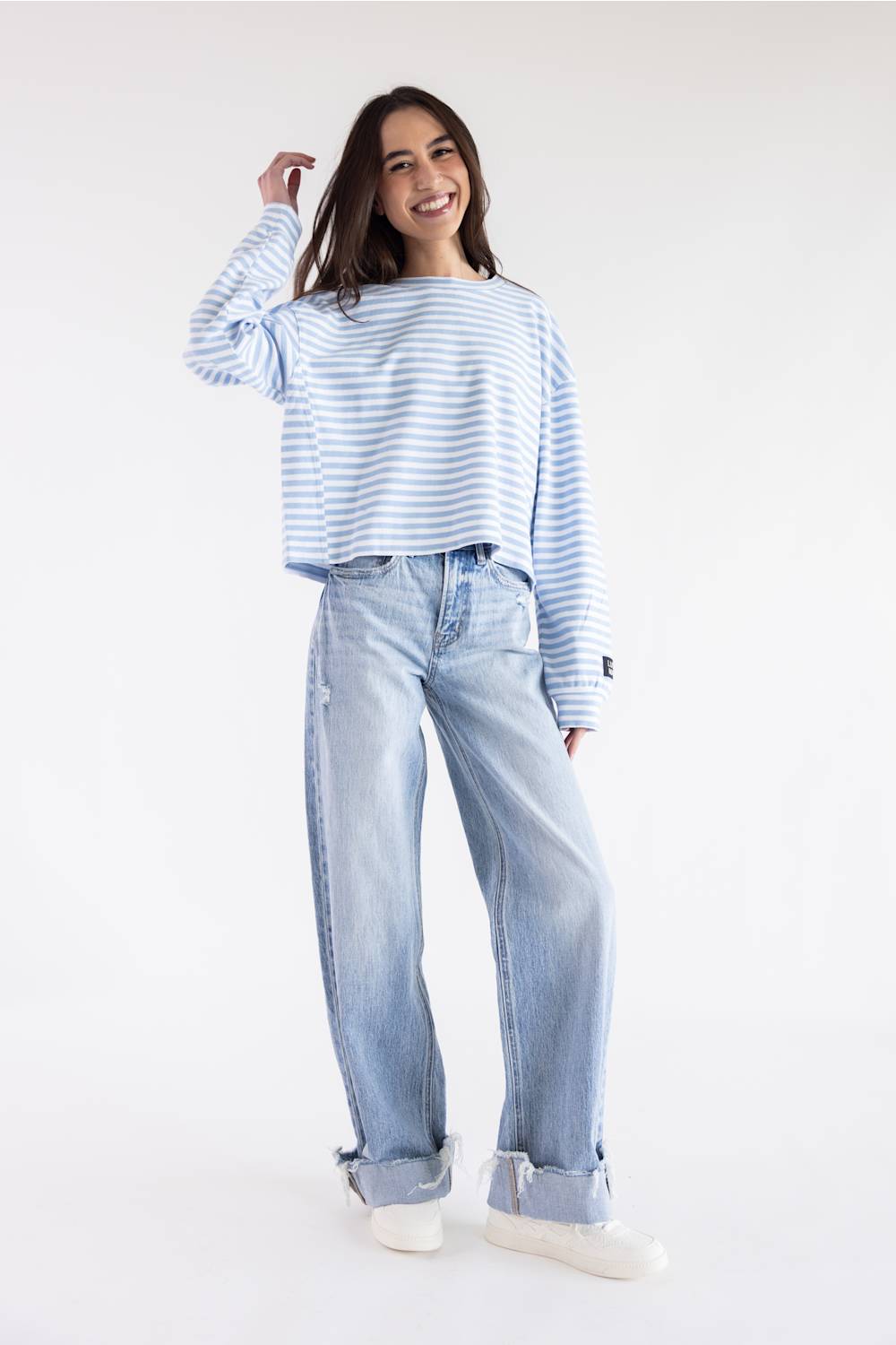 A woman modeling a light blue and white striped long-sleeve crop top with light-wash wide-leg jeans and white sneakers, standing against a plain white studio background.