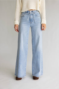 A model from the waist down wearing light-wash blue wide-leg jeans with a drawstring waist, paired with a cream ribbed sweater and brown shoes, against a light gray studio background.