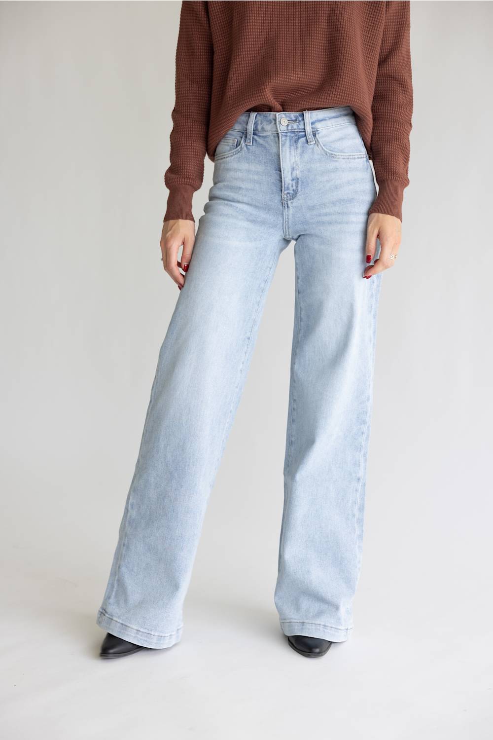 A model wearing light-wash wide-leg denim jeans, a brown ribbed knit sweater, and black pointed-toe heels, standing against a neutral light gray studio background.