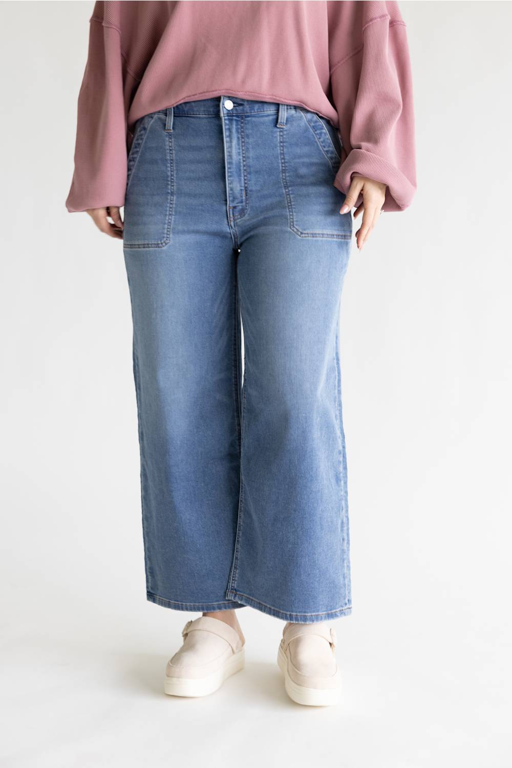 A person wearing light blue wide-leg denim jeans, a dusty pink long-sleeve top, and cream slip-on shoes, standing against a plain light gray studio background.