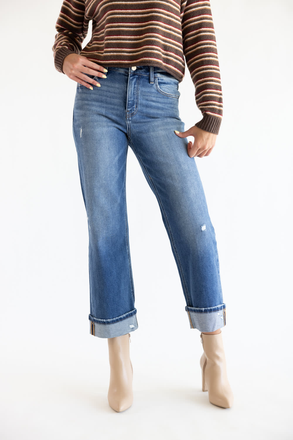 A model wearing a pair of medium-wash blue denim jeans with a straight-leg fit, cuffed hems, and subtle distressing, paired with a brown and beige striped sweater and beige ankle boots against a white studio background.