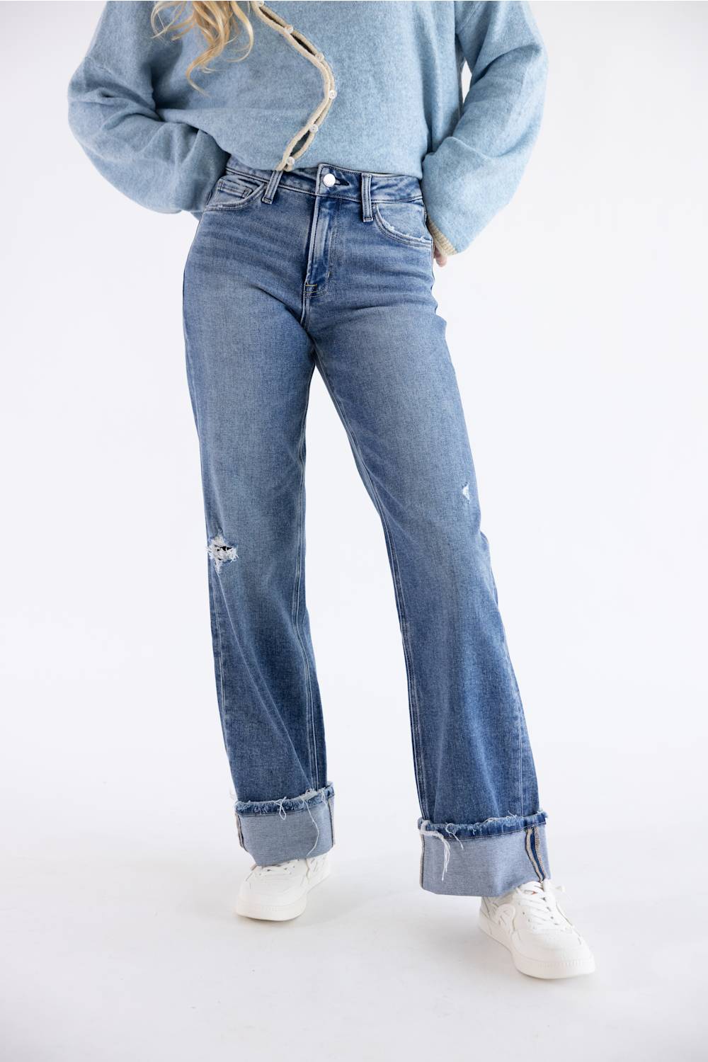 A person wearing light-wash blue distressed jeans with rolled cuffs, a light blue sweater, and white sneakers, photographed against a plain white studio background.