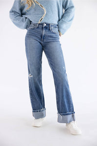 A person wearing light-wash blue distressed jeans with rolled cuffs, a light blue sweater, and white sneakers, photographed against a plain white studio background.