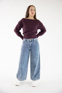 A woman modeling a dark purple off-the-shoulder knit sweater and light blue wide-leg jeans with small white star prints, paired with white sneakers, against a plain white studio background.