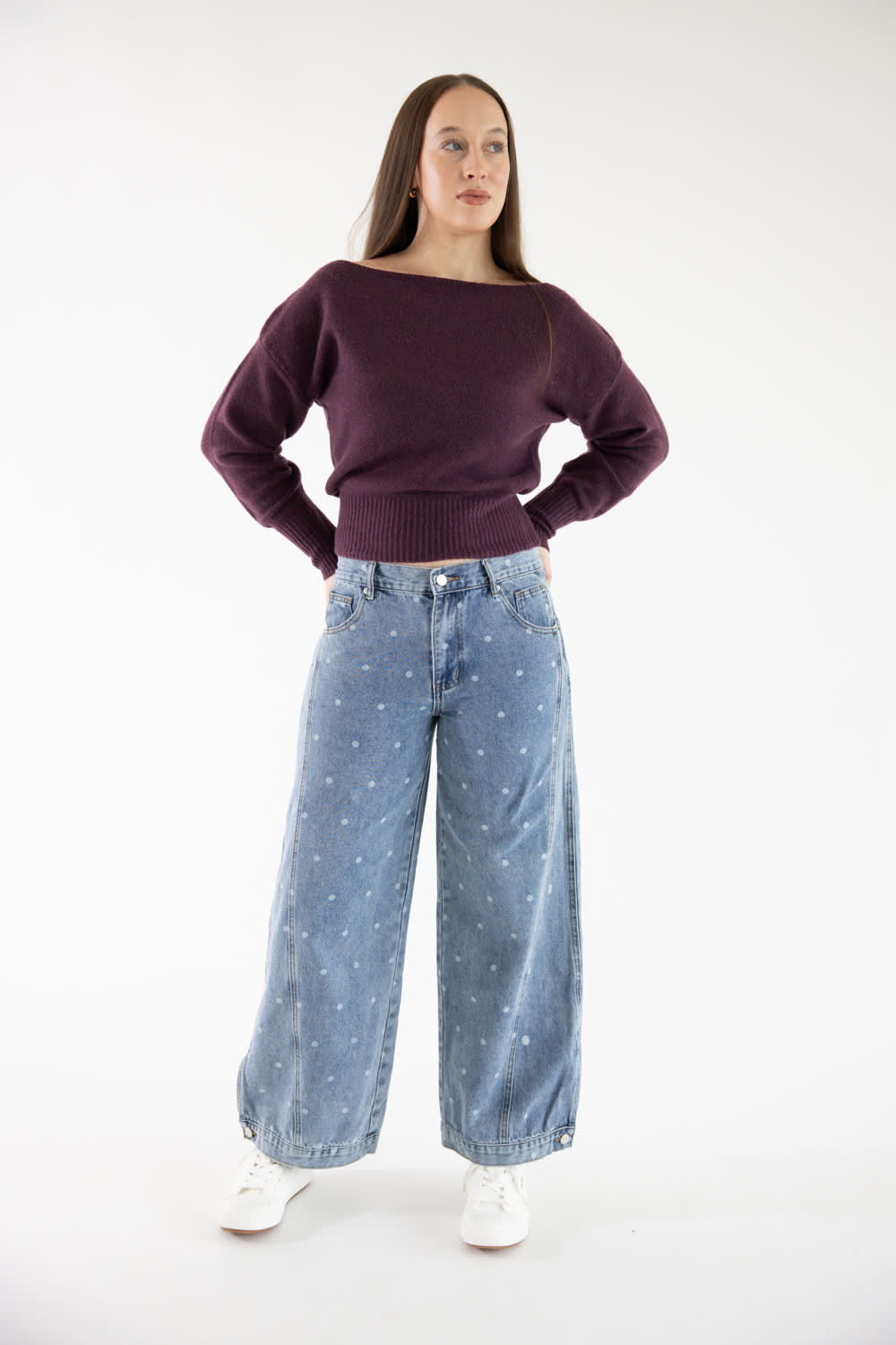 A woman modeling a dark purple off-the-shoulder knit sweater and light blue wide-leg jeans with small white star prints, paired with white sneakers, against a plain white studio background.