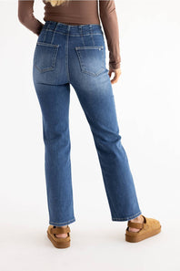 Rear view of a person wearing medium-wash blue jeans with a straight-leg fit, paired with a brown long-sleeve top and tan platform sandals, against a plain white studio background.