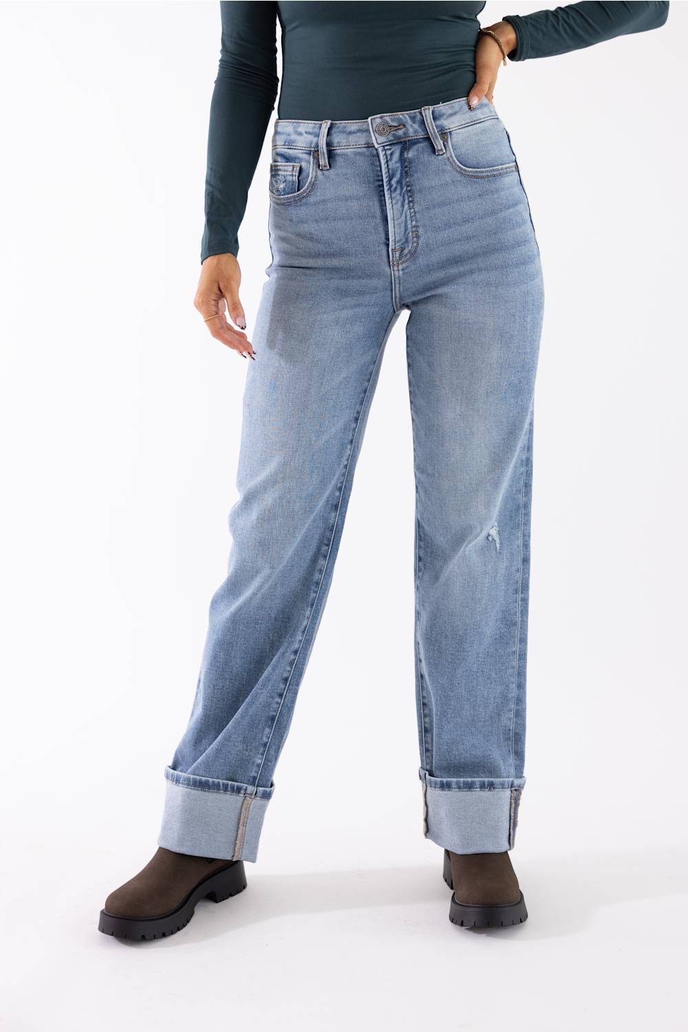 A person wearing light-wash blue straight-leg jeans with rolled cuffs, paired with a dark long-sleeve top and dark brown boots, photographed against a plain white studio background.