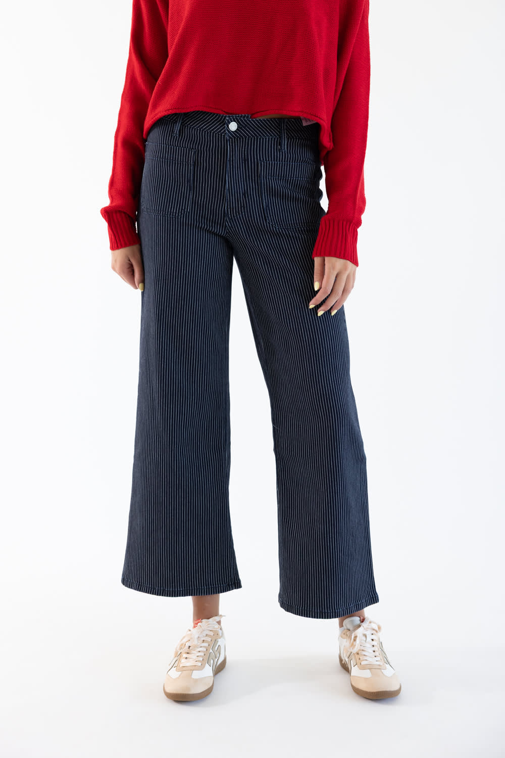 A model wearing dark navy blue pinstriped wide-leg cropped trousers, paired with a red sweater and beige sneakers, shown from the chest down on a white studio background.