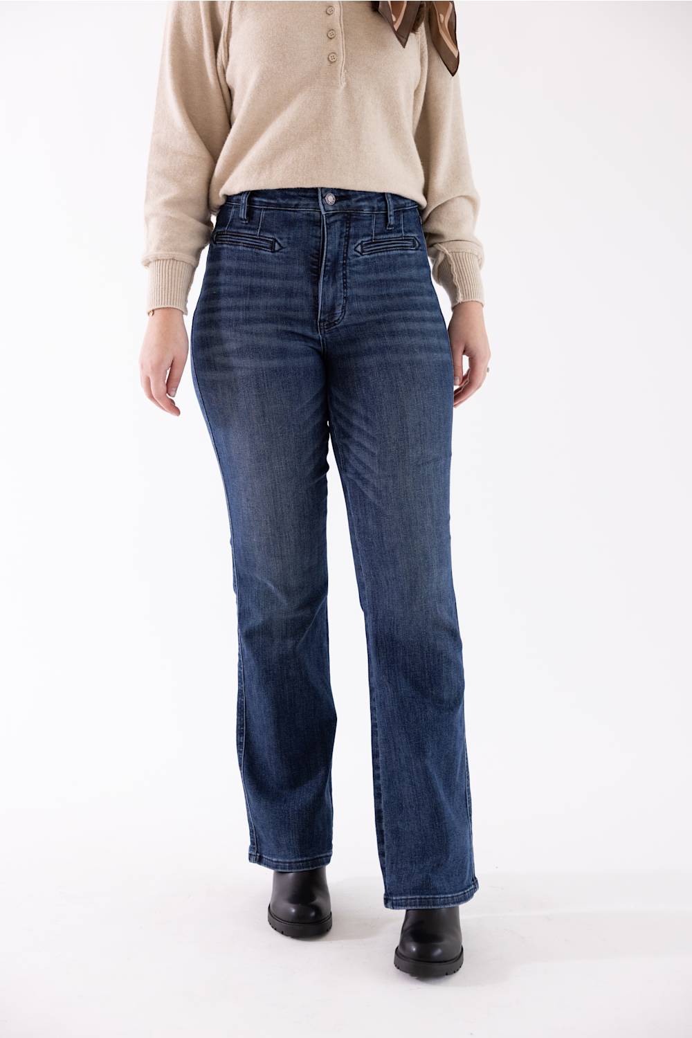 A model wearing a pair of dark blue straight-leg denim jeans, a beige long-sleeve button-up sweater, and black boots, standing against a plain white studio background.