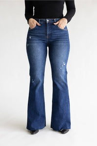 A person wearing dark blue high-rise flare jeans with distressed details and a black long-sleeve top, standing against a plain white studio background.