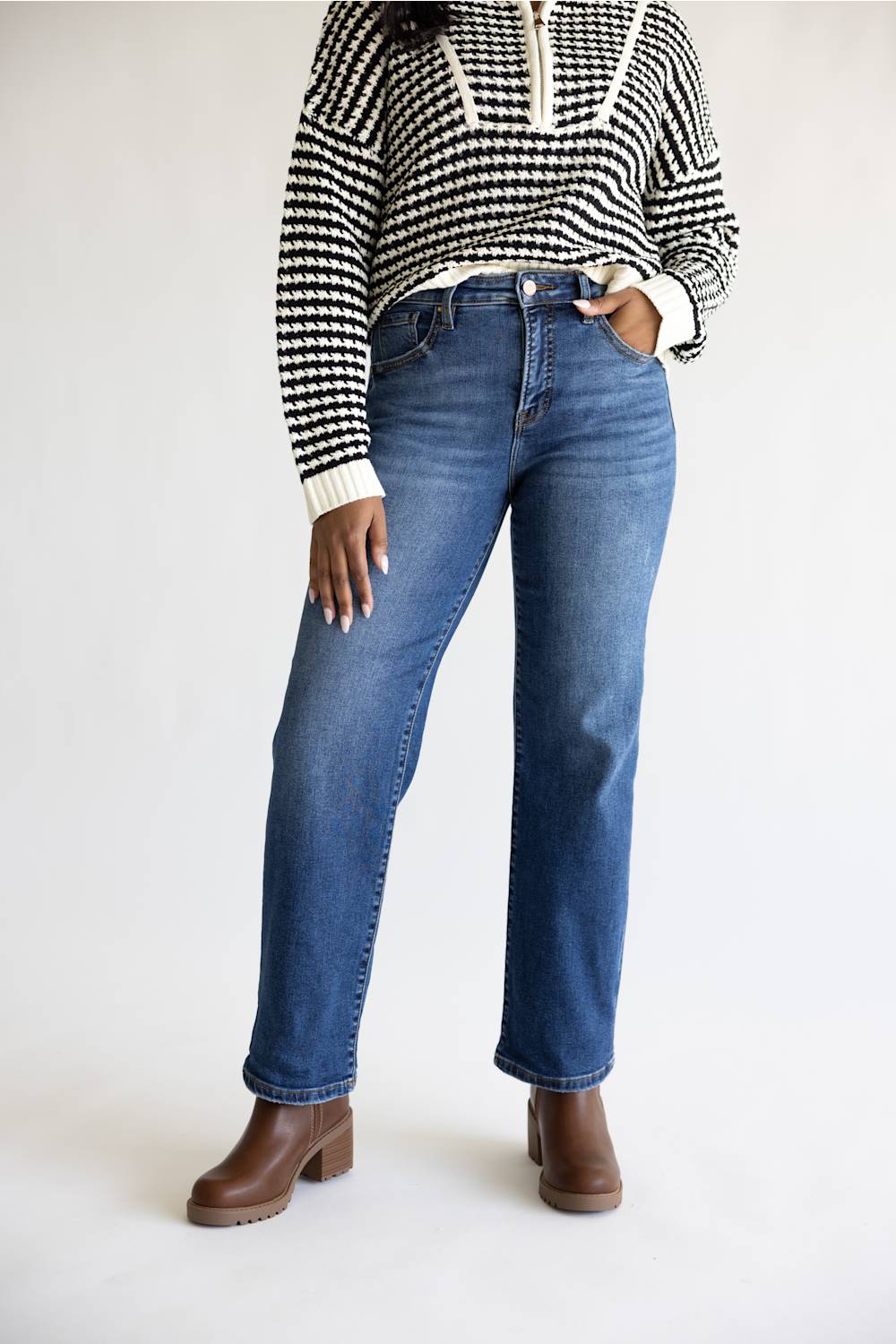 A model wearing a pair of medium-wash blue straight-leg jeans, paired with a black and white patterned knit sweater and brown leather boots, standing against a plain white studio background.