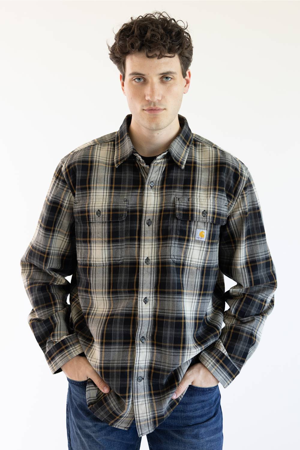 A man wearing a long-sleeve Carhartt plaid button-up shirt in black, gray, and white with thin orange stripes, paired with blue jeans, against a plain white background.