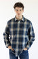 A man wearing a long-sleeve plaid button-up shirt in navy, gray, and beige with blue jeans, standing against a plain white background.