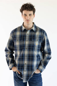 A man wearing a long-sleeve plaid button-up shirt in navy, gray, and beige with blue jeans, standing against a plain white background.