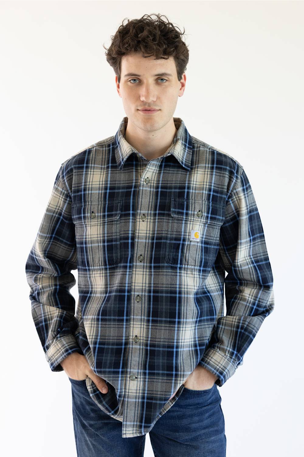 A man wearing a long-sleeve plaid button-up shirt in navy, gray, and beige with blue jeans, standing against a plain white background.