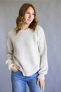 A woman wearing a light beige knit crewneck sweater with a scalloped ruffle trim, paired with blue jeans, standing against a neutral gray background.
