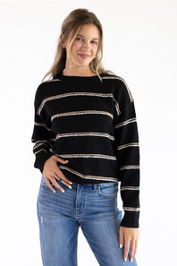 Model styled in a black crew neck knit sweater featuring textured tan and white horizontal stripes, ribbed hem and cuffs, paired with classic blue denim jeans against a white background.