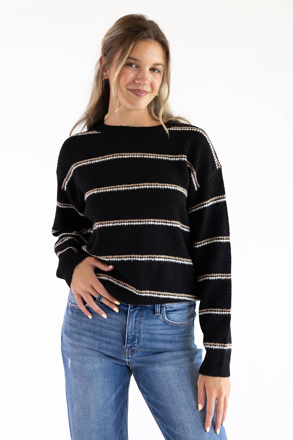 Model styled in a black crew neck knit sweater featuring textured tan and white horizontal stripes, ribbed hem and cuffs, paired with classic blue denim jeans against a white background.