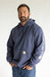 A man wearing a navy blue Carhartt hoodie with a white vertical logo on the sleeve and a small yellow logo patch on the chest pocket, paired with blue jeans against a plain white background.