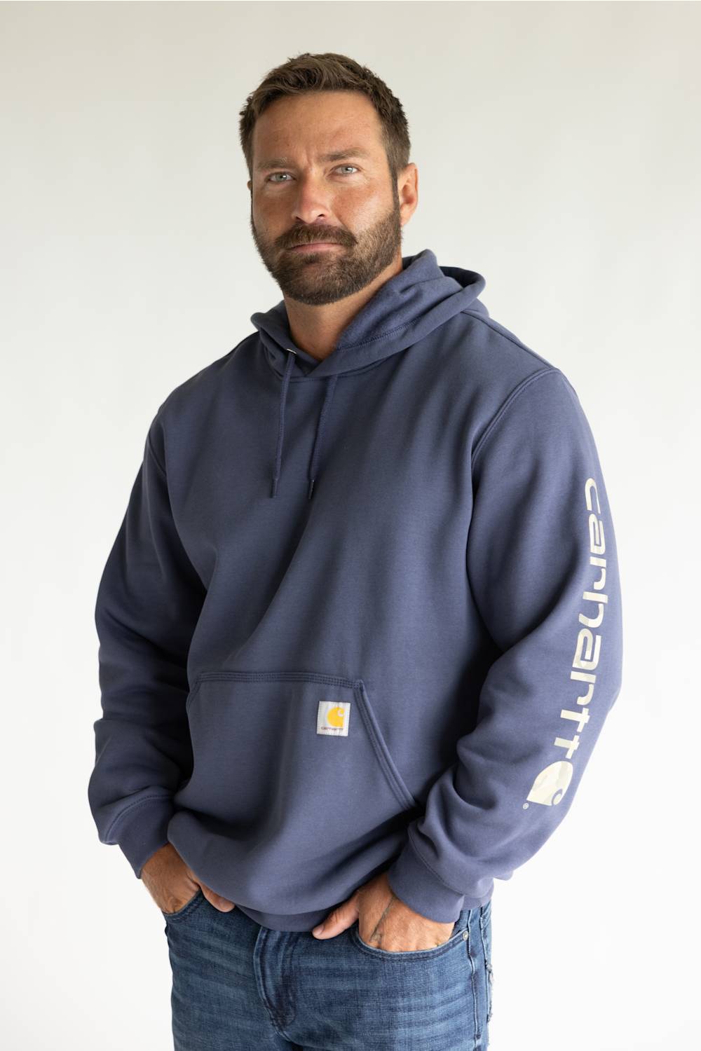 A man wearing a navy blue Carhartt hoodie with a white vertical logo on the sleeve and a small yellow logo patch on the chest pocket, paired with blue jeans against a plain white background.