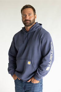 A man wearing a navy blue Carhartt hoodie with a white vertical logo on the sleeve and a small yellow logo patch on the chest pocket, paired with blue jeans against a plain white background.