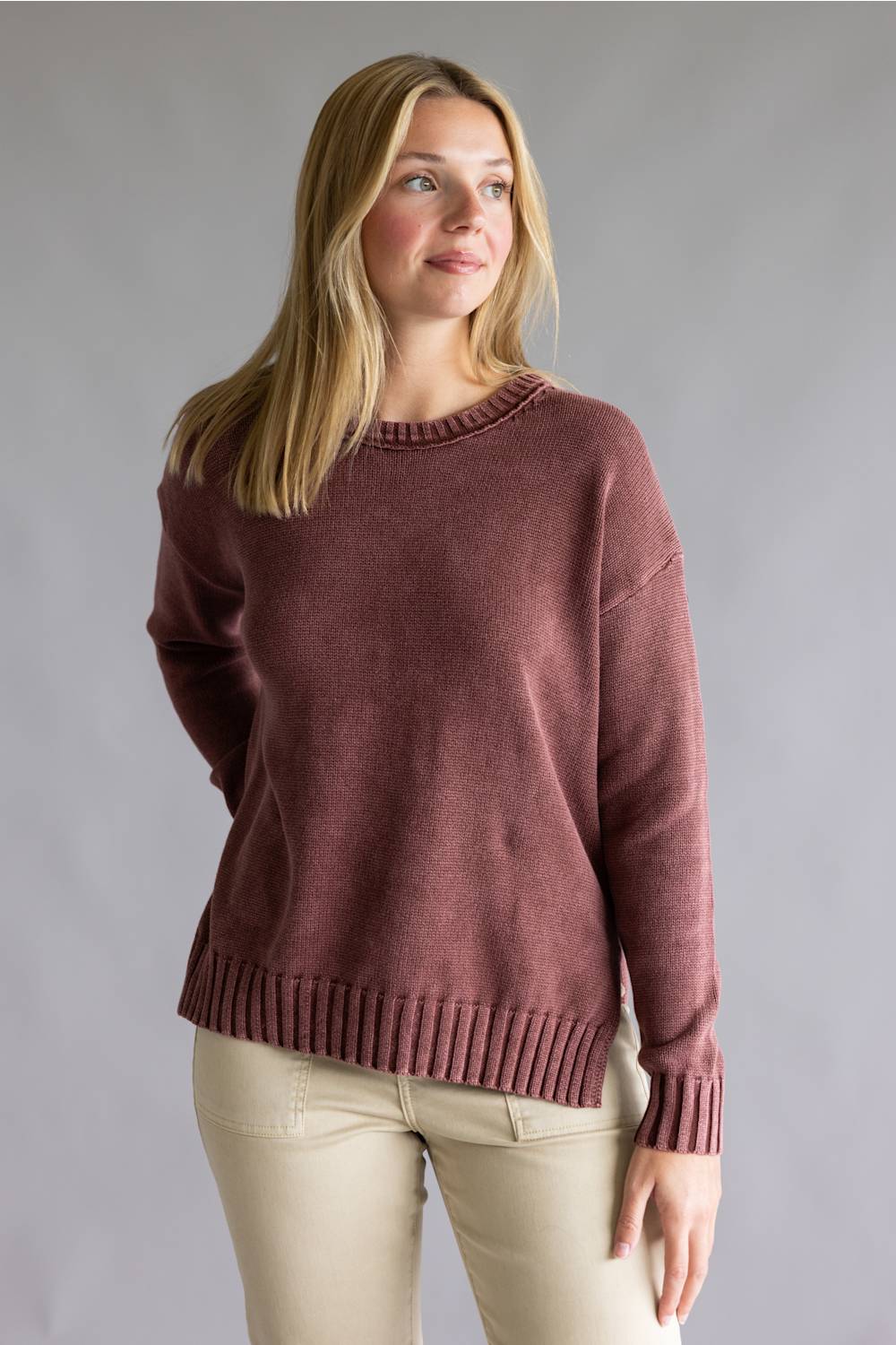 A woman wearing a dusty rose knit sweater with ribbed collar, cuffs, and hem, paired with beige pants, photographed against a neutral gray studio background.