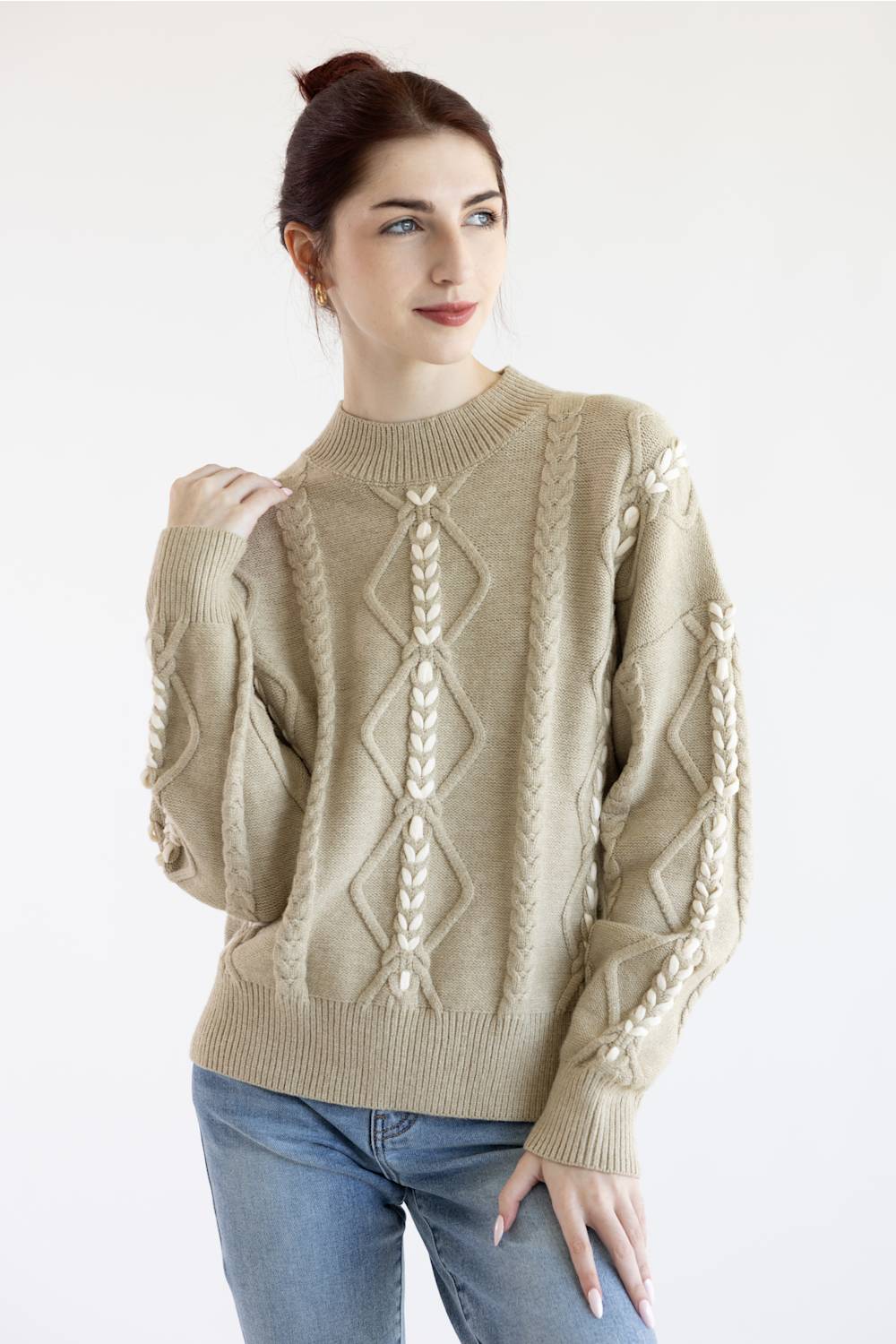 A woman wearing a beige cable-knit sweater with cream-colored diamond and leaf patterns, paired with blue jeans, against a plain white background.
