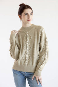 A woman wearing a beige cable-knit sweater with cream-colored diamond and leaf patterns, paired with blue jeans, against a plain white background.