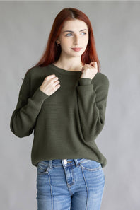 A woman with red hair wearing an olive green waffle-knit long-sleeve sweater and blue jeans, posing against a neutral gray studio background.