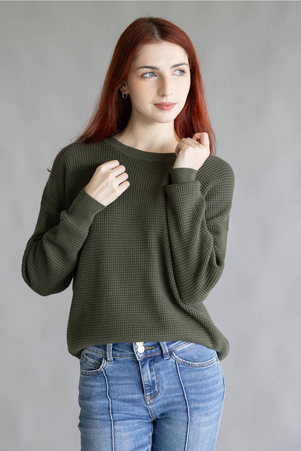 A woman with red hair wearing an olive green waffle-knit long-sleeve sweater and blue jeans, posing against a neutral gray studio background.