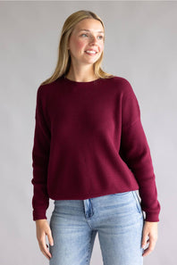 A woman wearing a textured waffle-knit crewneck sweater in deep burgundy, paired with light blue jeans, standing against a neutral gray studio background.