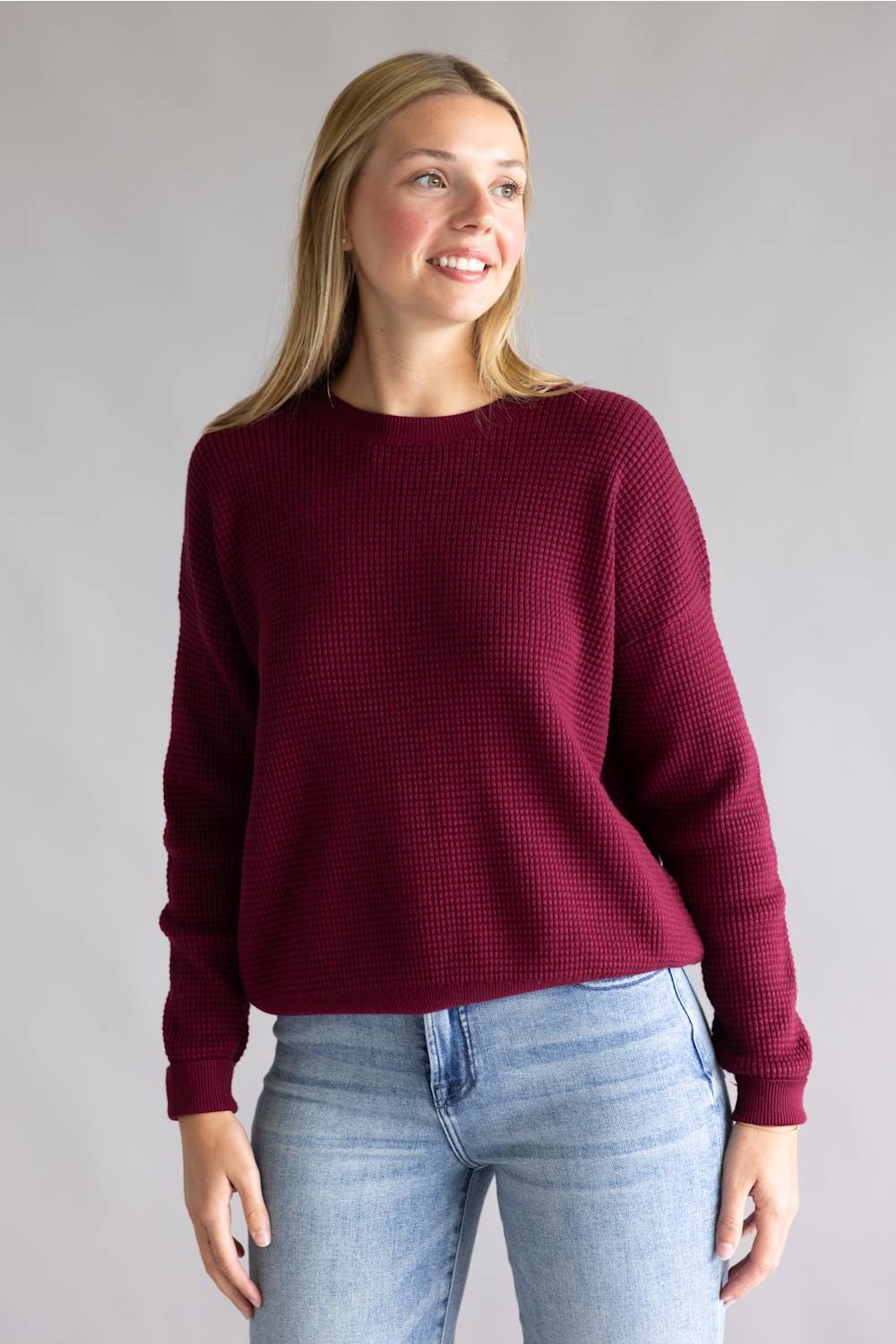 A woman wearing a textured waffle-knit crewneck sweater in deep burgundy, paired with light blue jeans, standing against a neutral gray studio background.