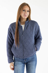 A woman modeling a slate blue diamond-quilted zip-up fleece jacket, paired with a white top and blue jeans, against a plain white studio background.